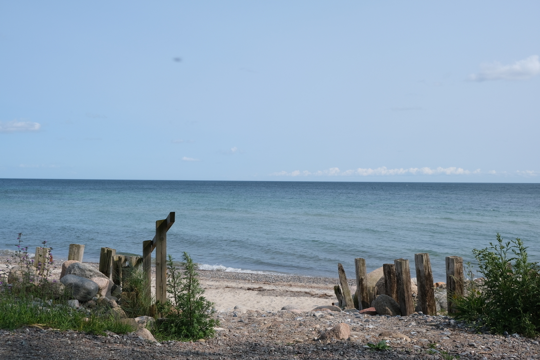 A calm beach scene with clear blue water, sandy shore, and wooden posts lining the coastline.
