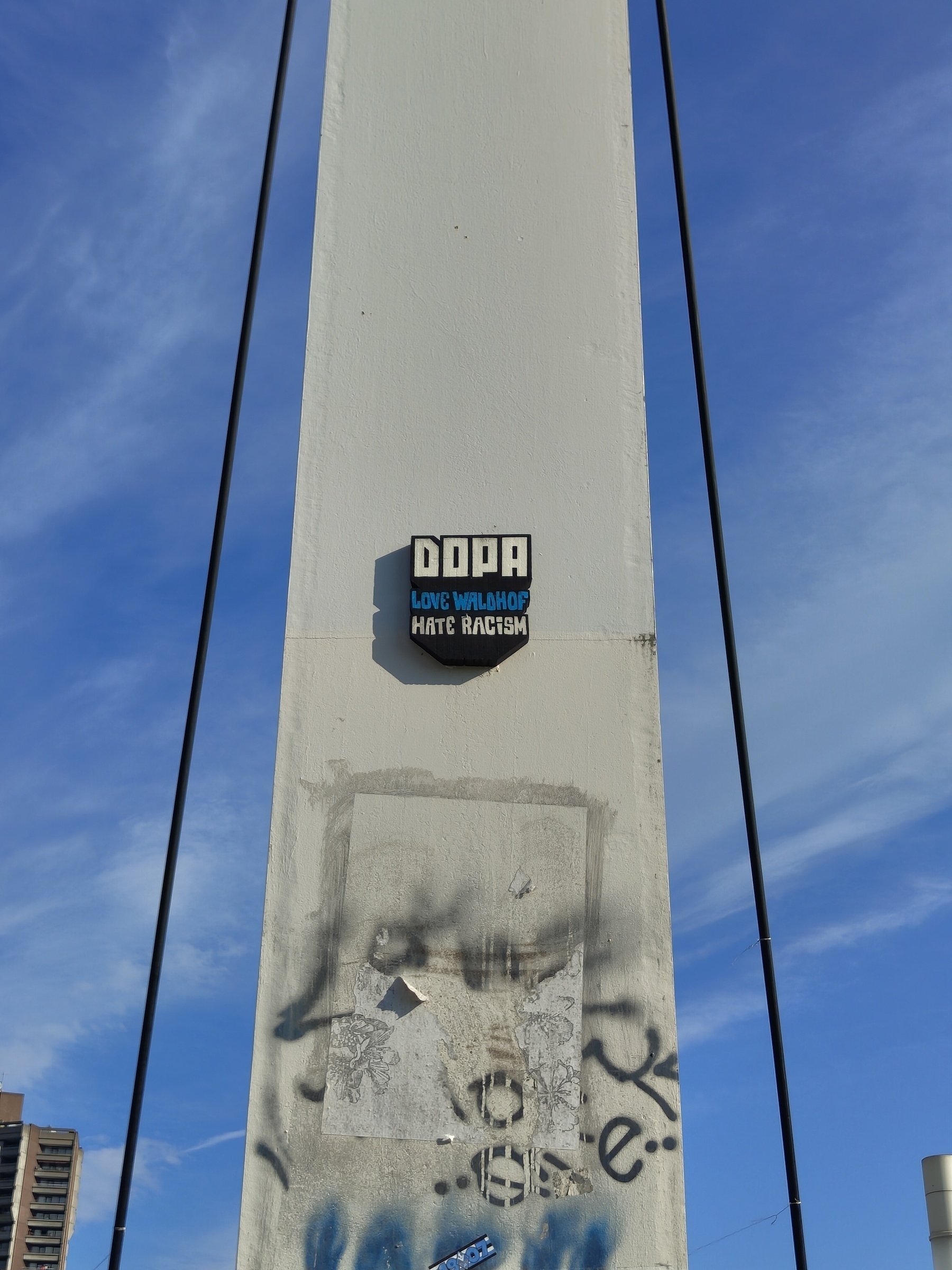 A tall, concrete structure features a sticker or plaque that says LOVE WALADOO HATE RACISM against a blue sky background.