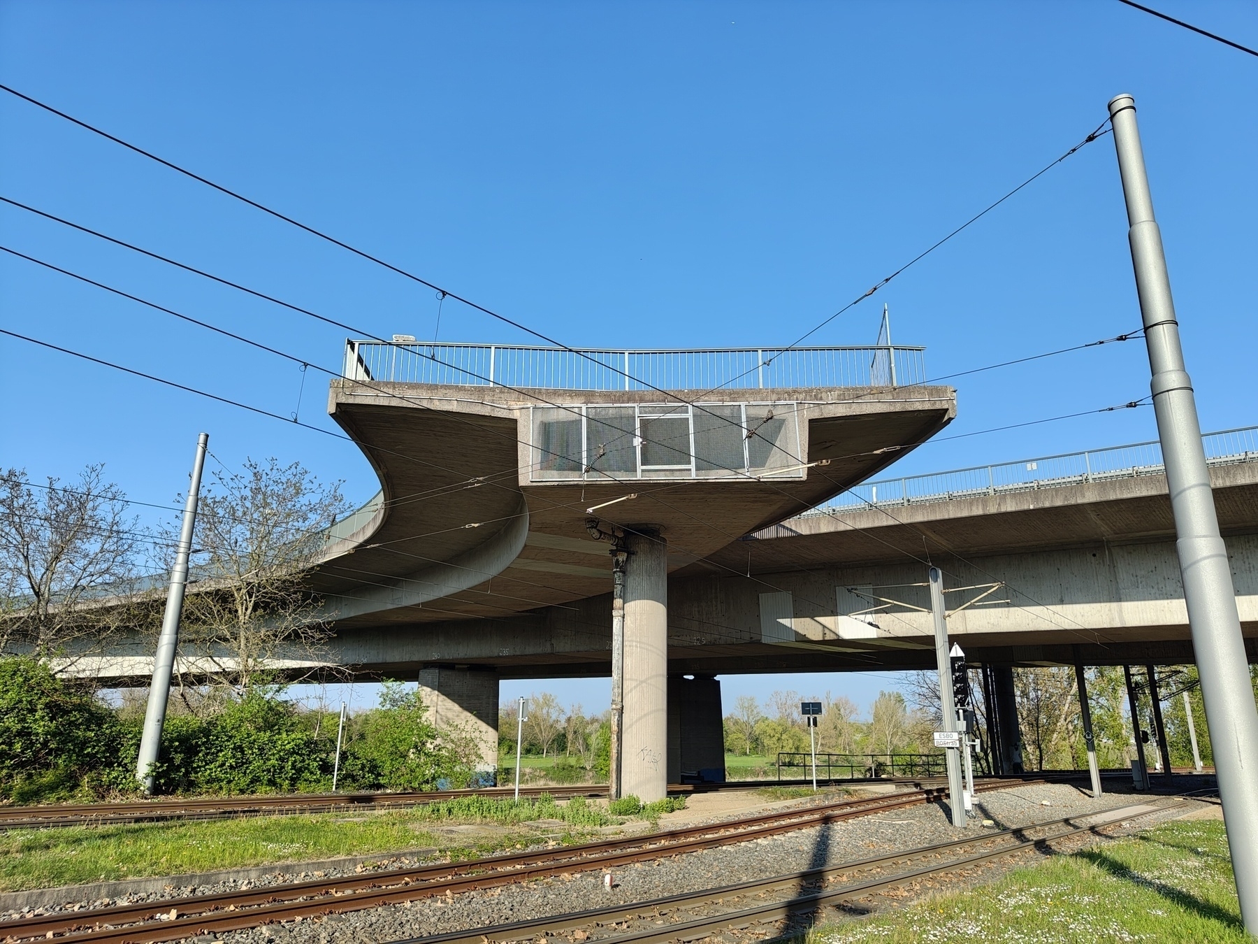 A concrete overpass with a distinct triangular section is situated above railway tracks against a clear blue sky.