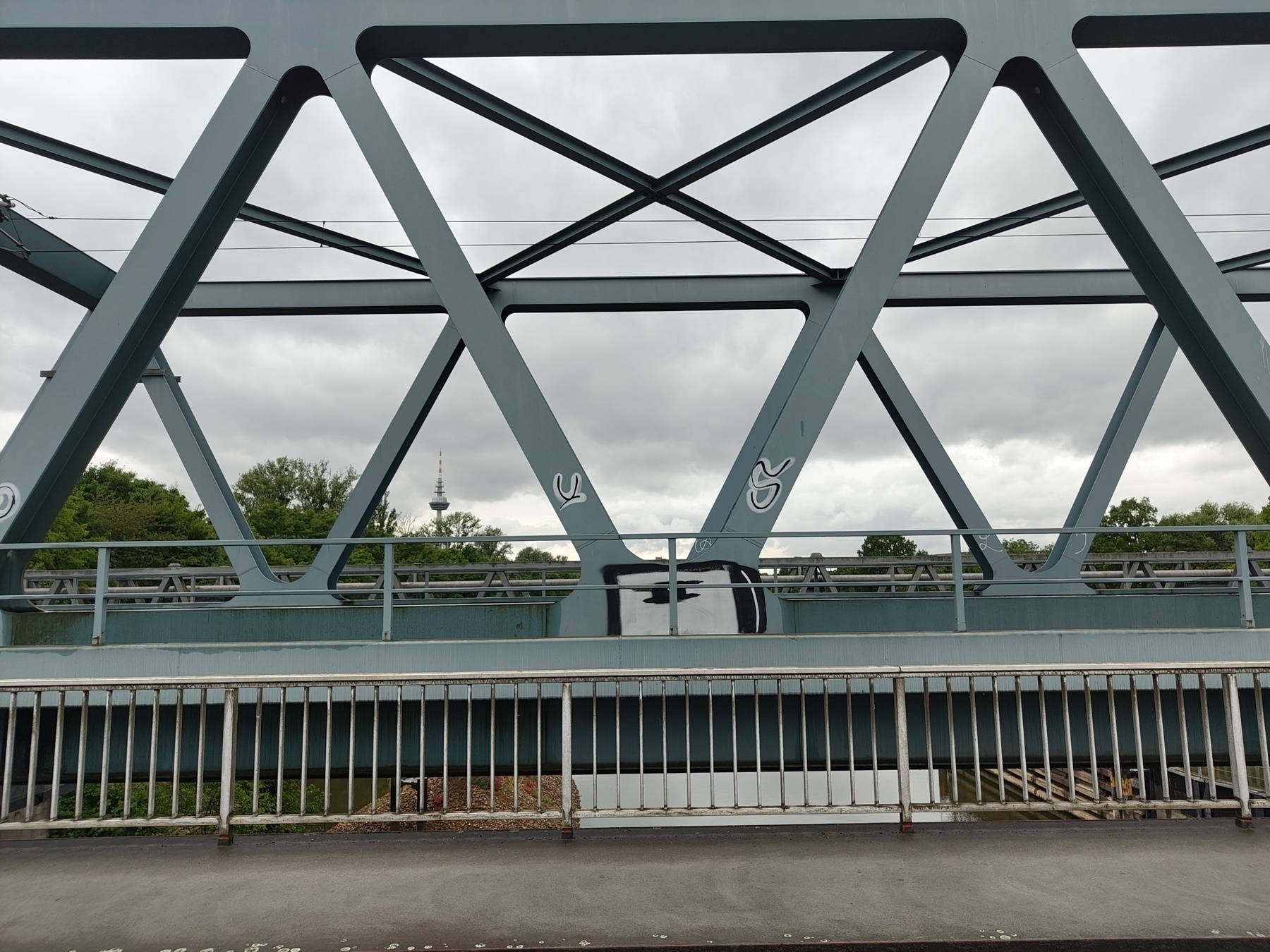 A metal bridge with a crisscross design stands against a cloudy sky, with greenery visible in the background.