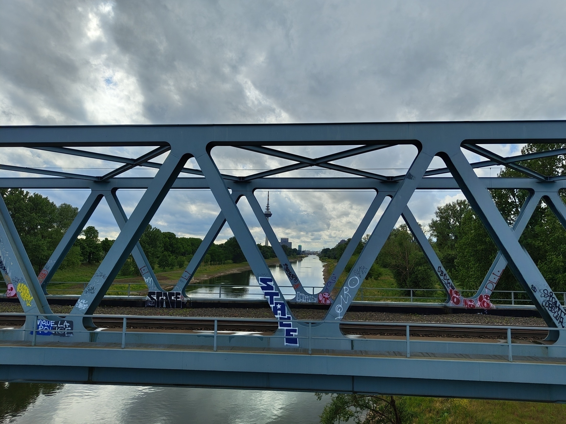 A metal bridge with graffiti spans over a river, with trees and clouds in the background.