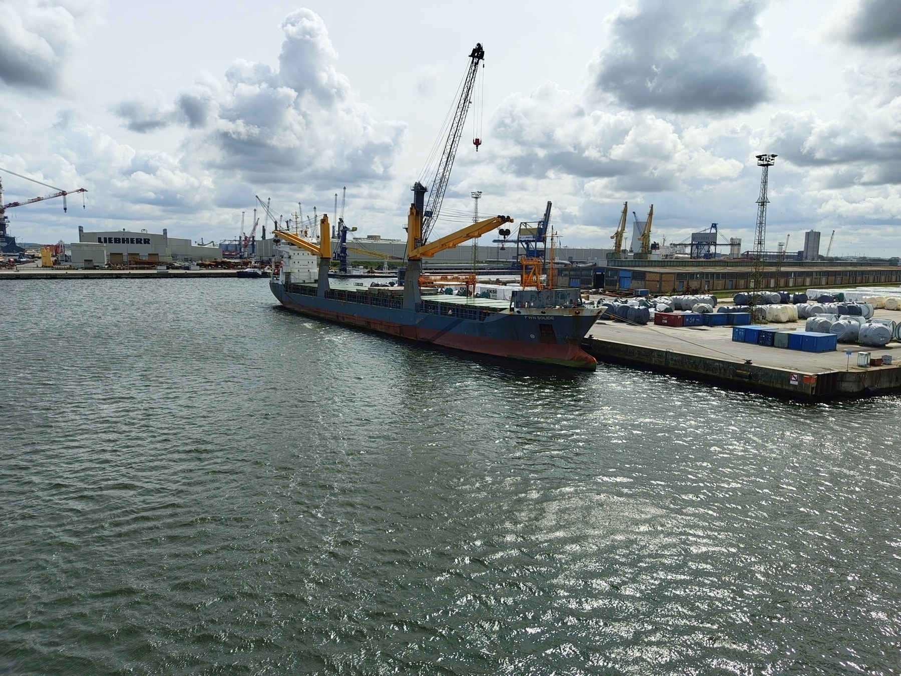 A cargo ship is docked at an industrial port with cranes and warehouses in the background.