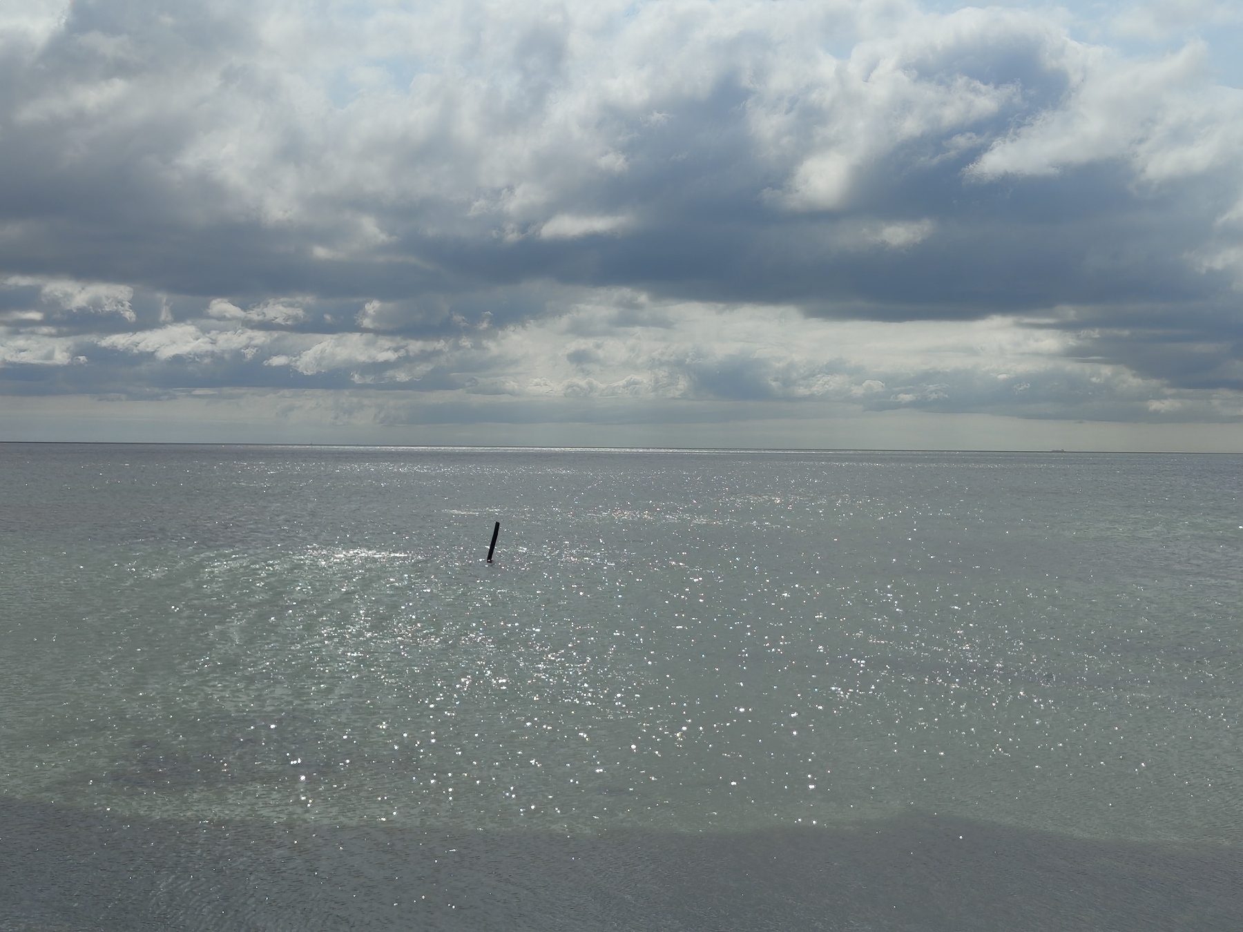 A calm sea stretches to the horizon under a cloudy sky, with sunlight reflecting on the water's surface and a single pole emerging from the ocean.
