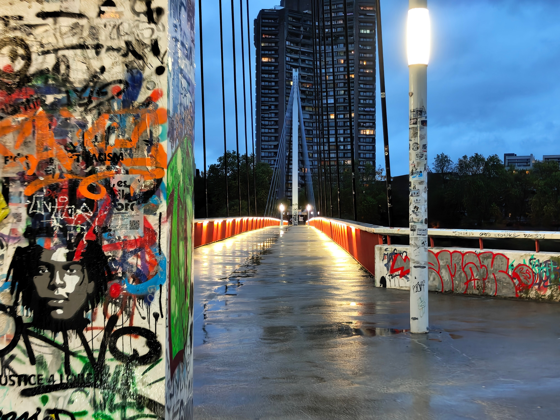 A graffiti-covered urban bridge with bright lights leads toward a tall building against a dusk sky.